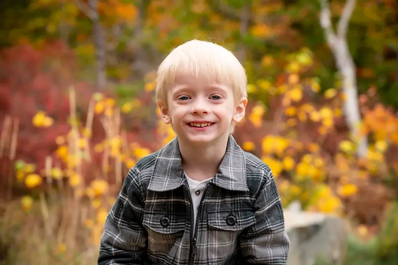 A young boy with blond hair smiles during a fall photo session at Moments in Time Photography Studio, surrounded by vibrant autumn leaves and warm natural colors.