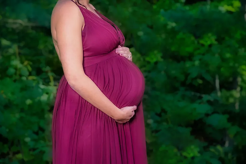 Pregnant mother in a sleeveless burgundy maternity dress holding her bump outdoors during a Halifax maternity photography session.