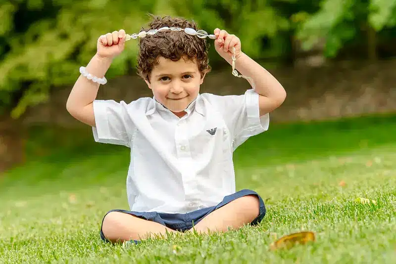 Playful outdoor portrait of a young curly-haired boy sitting cross-legged on green grass by Moments in Time Photography Studio Halifax